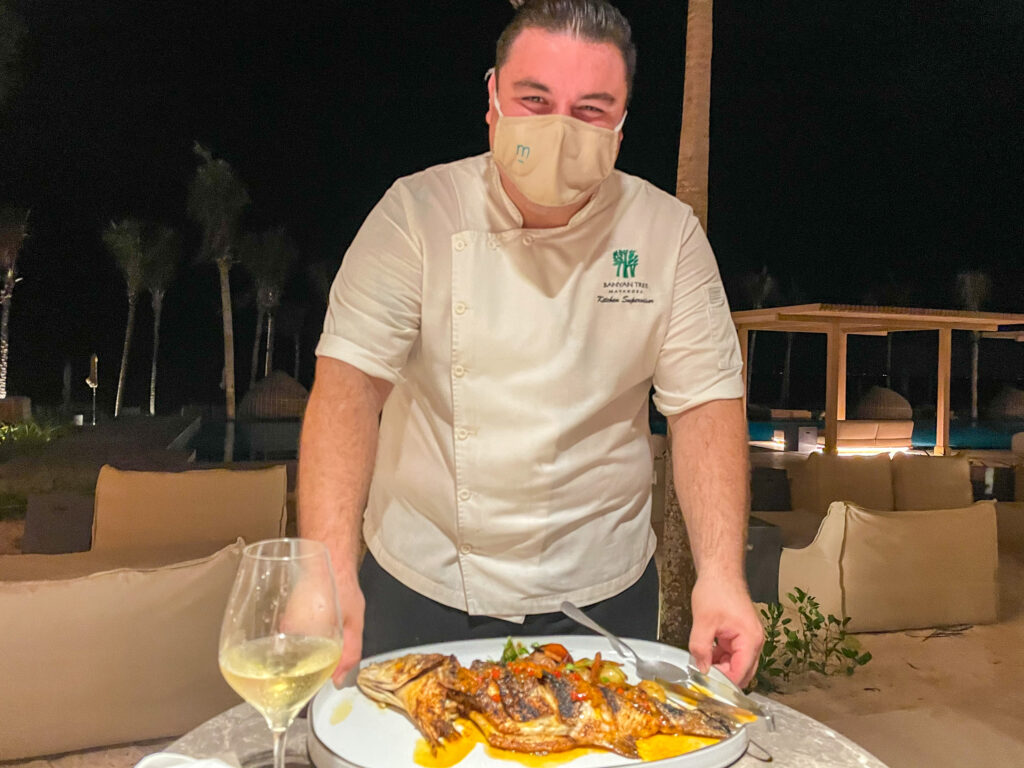 A chef presenting a whole grilled fish on a platter beside the lit pool after dark