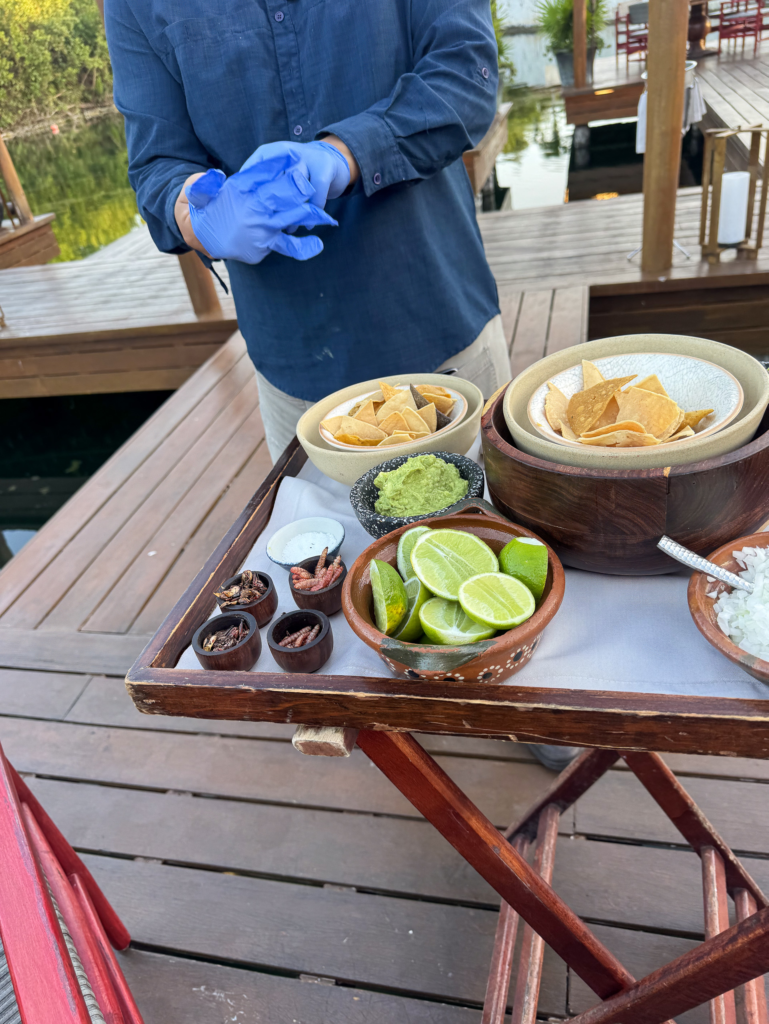 A waiter in blue gloves preparing guacamole at a wooden trolley of chips, limes and salsas on the deck