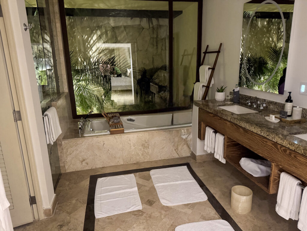A bathroom with a sunken tub beside a garden window, stone-topped double vanity and rolled white towels