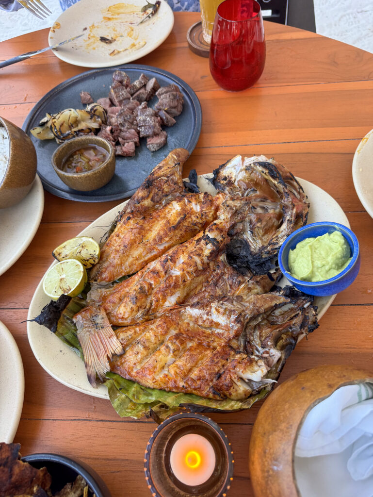 A whole grilled fish with charred lime, guacamole and grilled steak on a wooden table