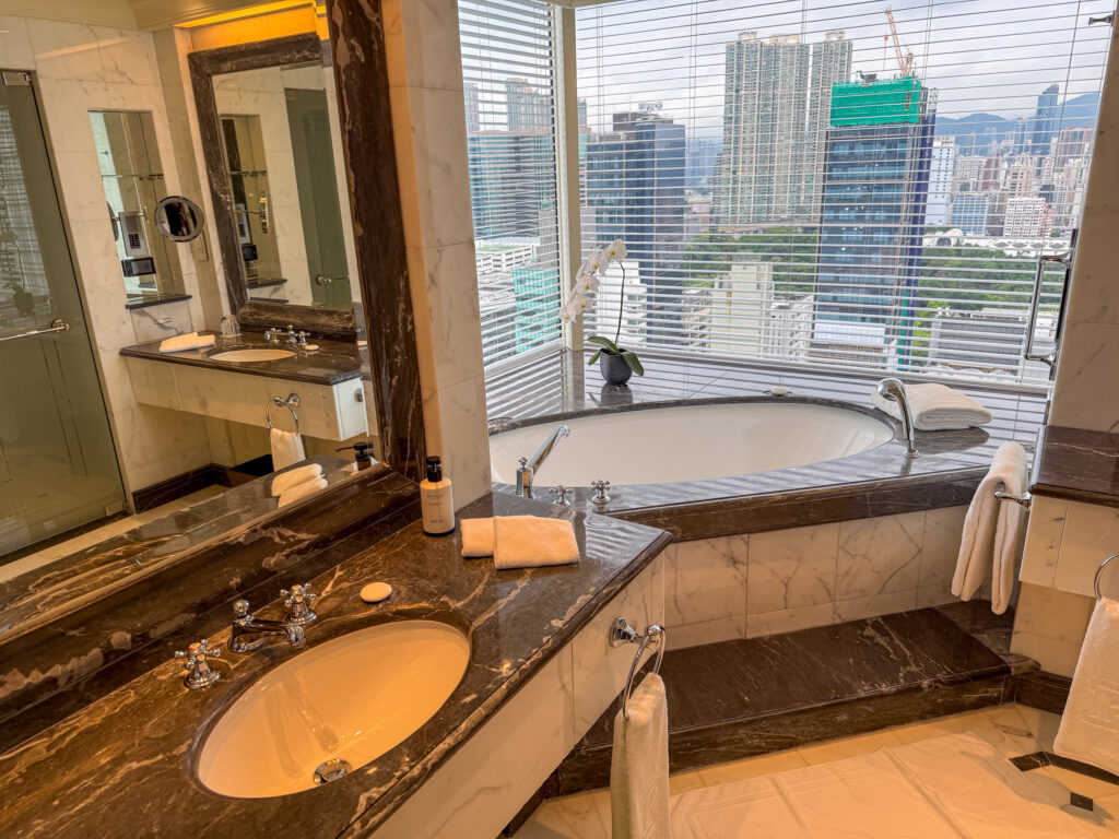 A marble bathroom with twin basins, a mirrored wall and a tub under the window with a white orchid on the sill