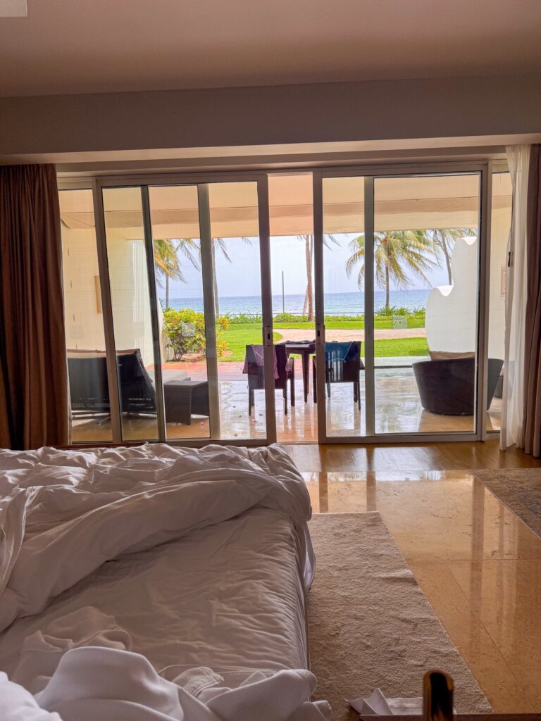 Rumpled white bed and marble floor facing glass doors open to palms, lawn and sea