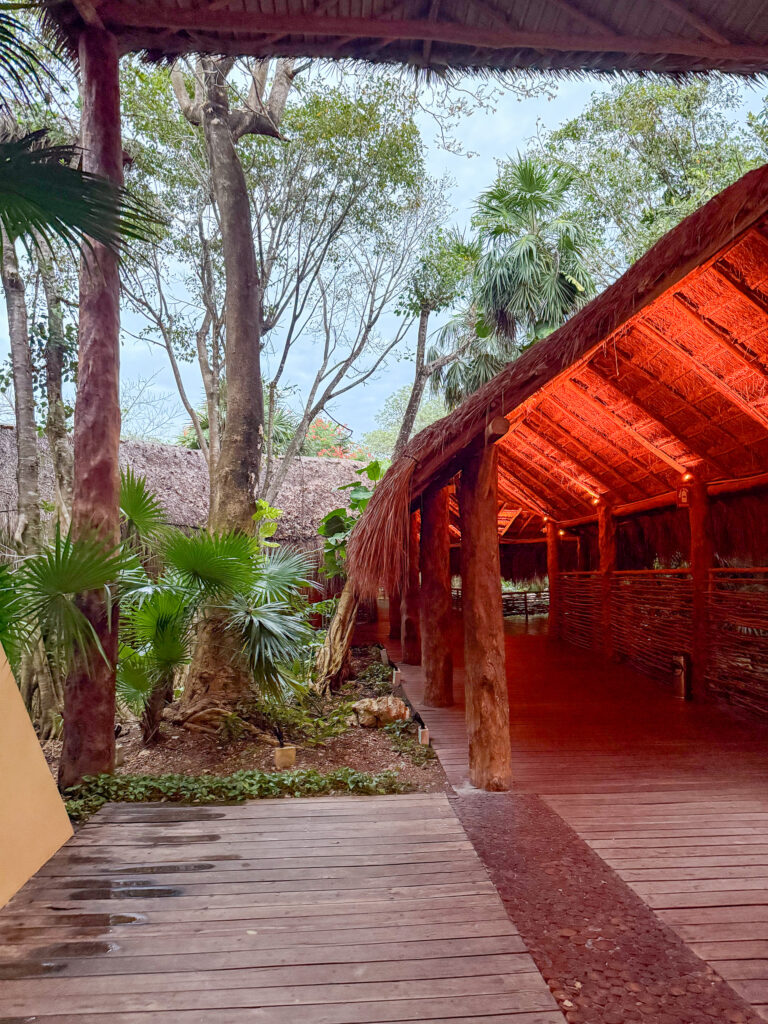 Thatched open walkway lit warm orange, running on rough-hewn posts through palms and jungle