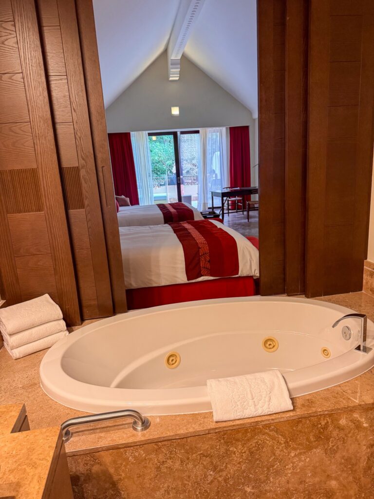 Oval whirlpool tub in a travertine bathroom looking through to a bed with red runner