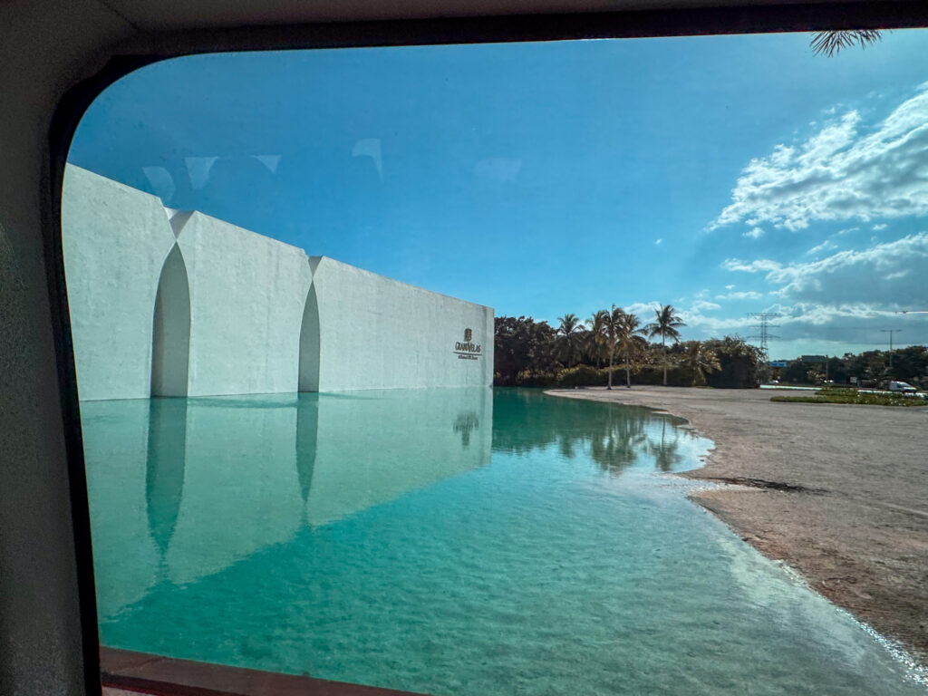 Curved white wall reflected in a shallow turquoise arrival pool, seen through a vehicle window