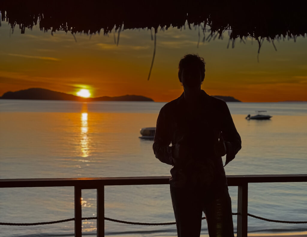 J.C. Mercer silhouetted against a sunset over the water, giving a thumbs up from a beachfront deck
