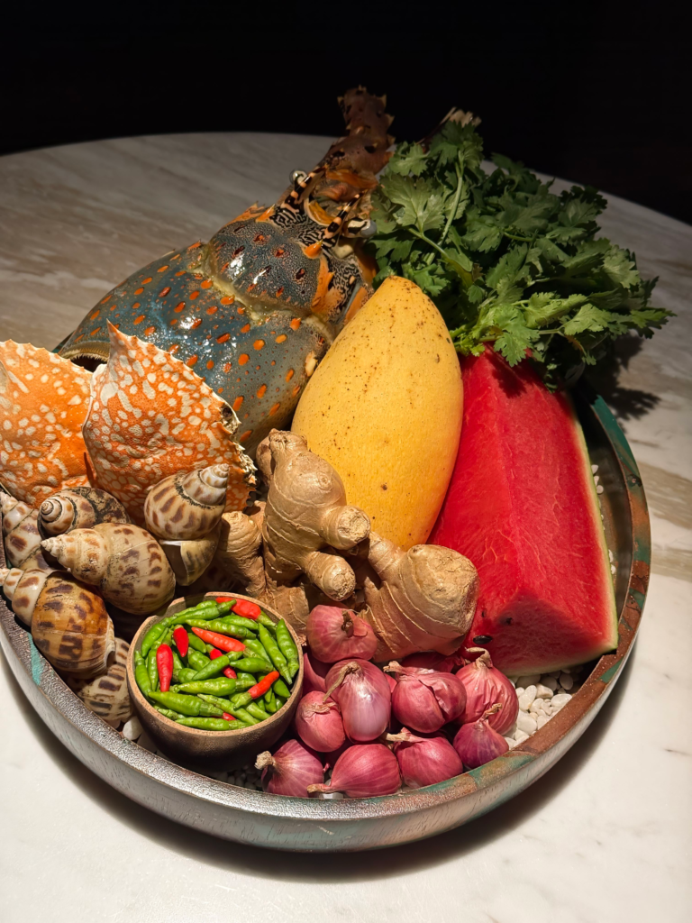 A shallow copper-rimmed tray of raw southern Thai ingredients on white pebbles