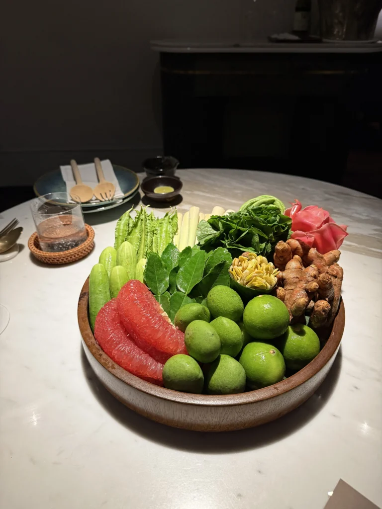 A wooden bowl of raw southern Thai produce: winged beans, cucumbers, baby corn, betel leaves, torch ginger flower, fresh turmeric, pomelo and green mangoes