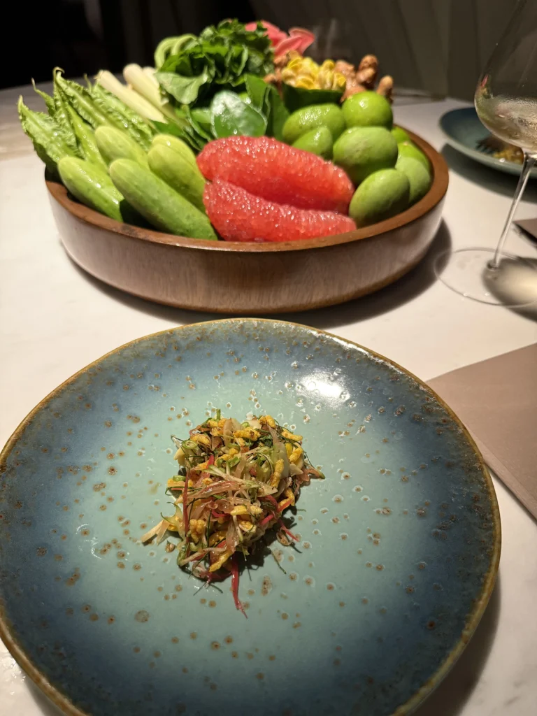 A small herb and pomelo salad heaped on a speckled blue plate, the bowl of raw produce behind it