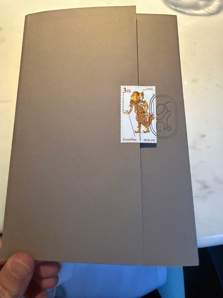 The closed menu, a grey card folder sealed with a Thai postage stamp, held above the marble table