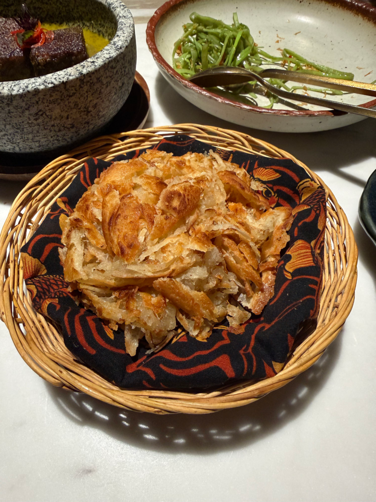 Torn layers of flaky roti in a cloth-lined basket beside a curry and a bowl of greens
