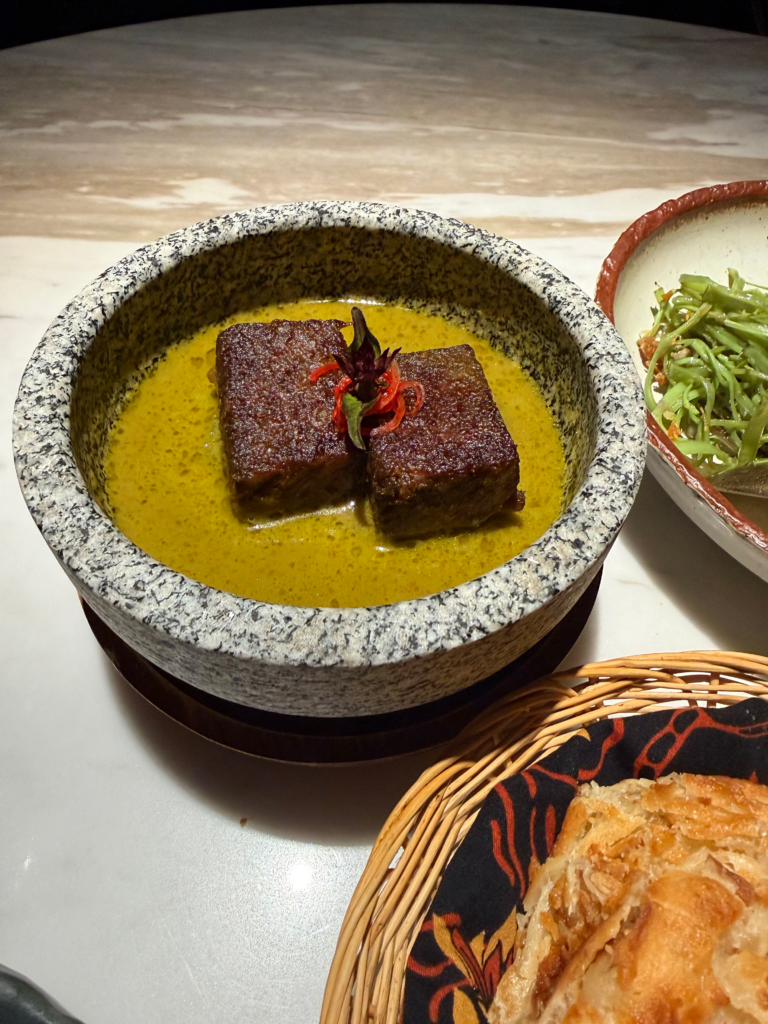 Two blocks of dark braised meat in a yellow curry served in a granite mortar, a basket of roti alongside