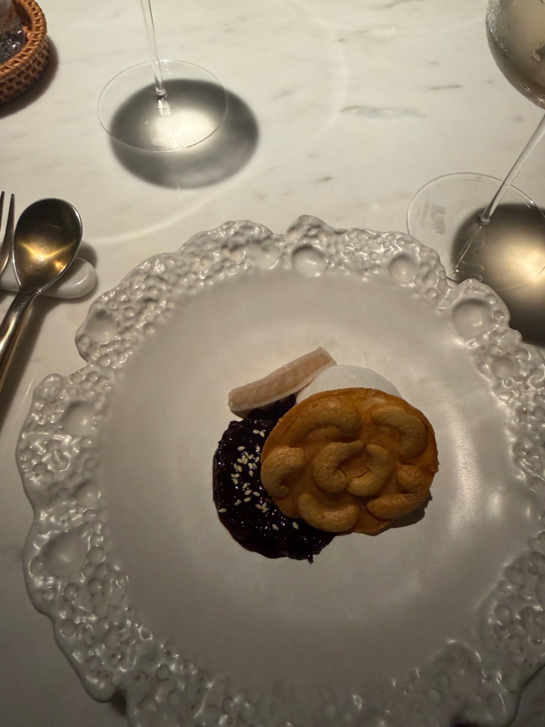 A dessert of black sticky rice with sesame, coconut ice cream and a cashew-studded wafer on a textured white plate