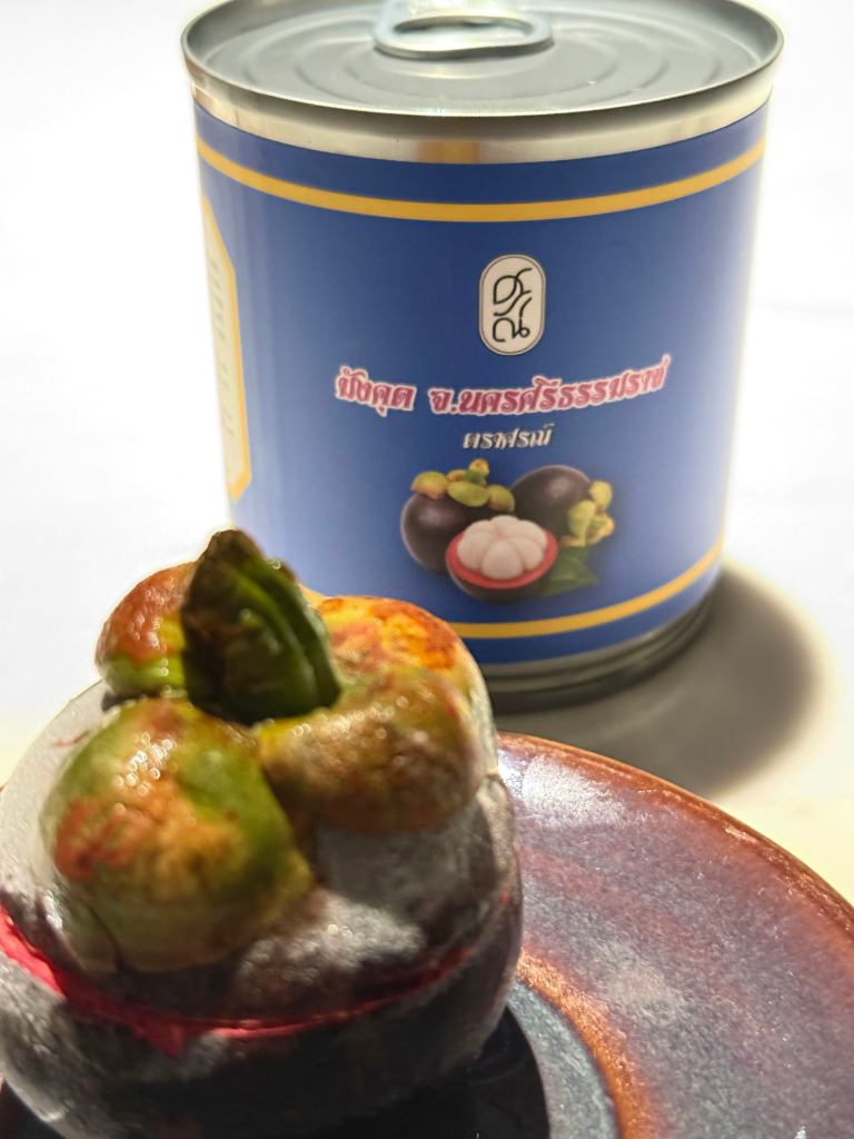 A whole mangosteen dessert on a glazed plate in front of a Thai tin of canned mangosteen