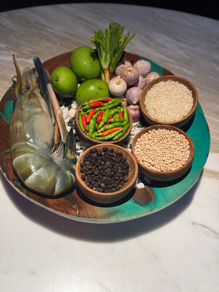 A ceramic tray of raw ingredients: a whole river prawn, green mangoes, garlic, bird's eye chillies, jasmine rice and black and white peppercorns