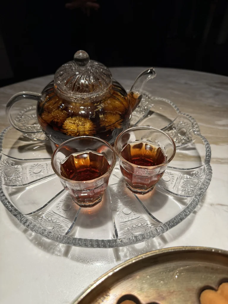 A glass teapot of flowering chrysanthemum tea and two amber-filled cups on an etched glass tray