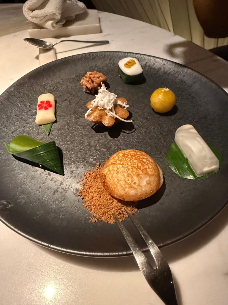 Seven Thai petits fours arranged on a dark slate plate, a coconut crisp sitting on a heap of palm sugar