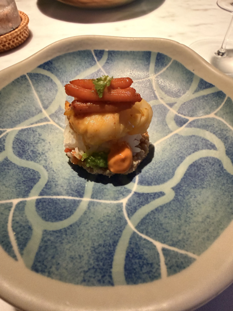 A single bite of jasmine rice topped with shellfish and glazed red strips, on a blue and white glazed plate