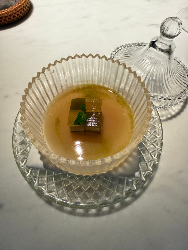 A clear consomme in a fluted glass bowl, two cubes and a mint leaf at its centre, the glass lid set aside
