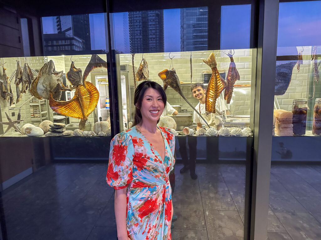 A diner standing in front of the glass-fronted dry-ageing room, whole fish hanging on hooks behind her