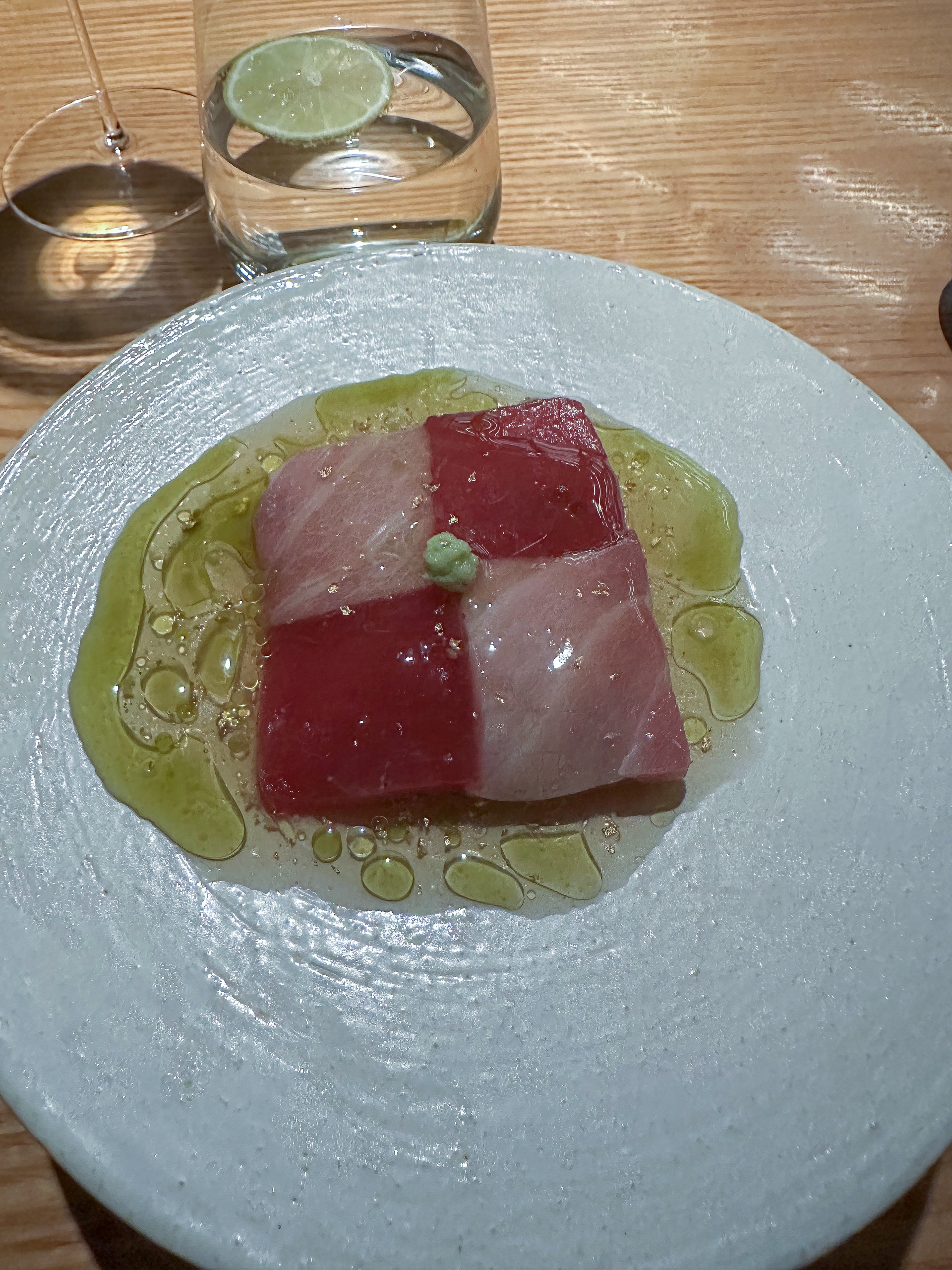 Checkerboard slices of tuna loin and belly in green olive oil on a pale ceramic plate, a glass of water alongside