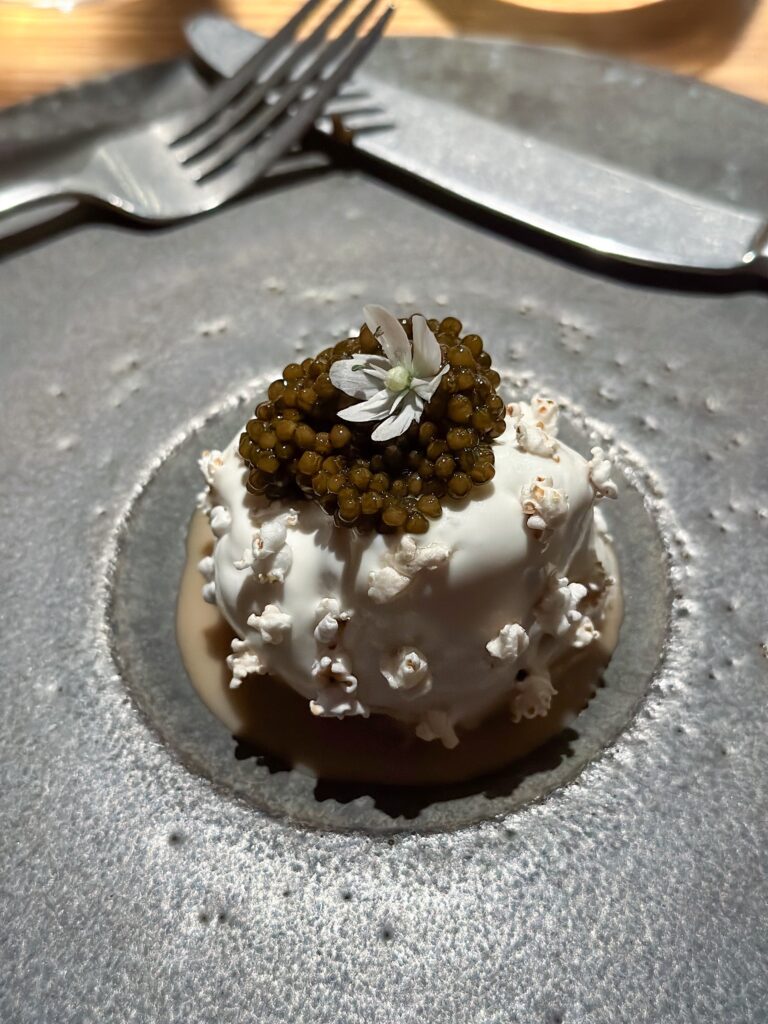 A quenelle of cream scattered with popcorn and crowned with caviar and a white flower, set in the hollow of a stone plate