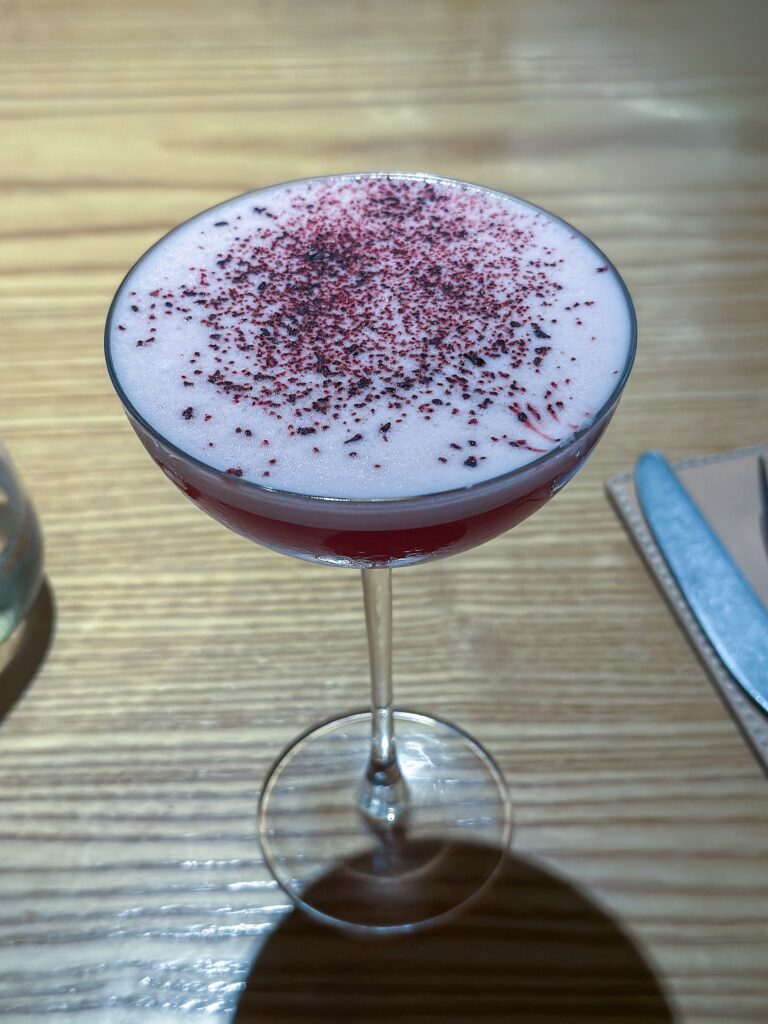 A coupe of deep red cocktail under a white foam cap dusted with dried berry powder