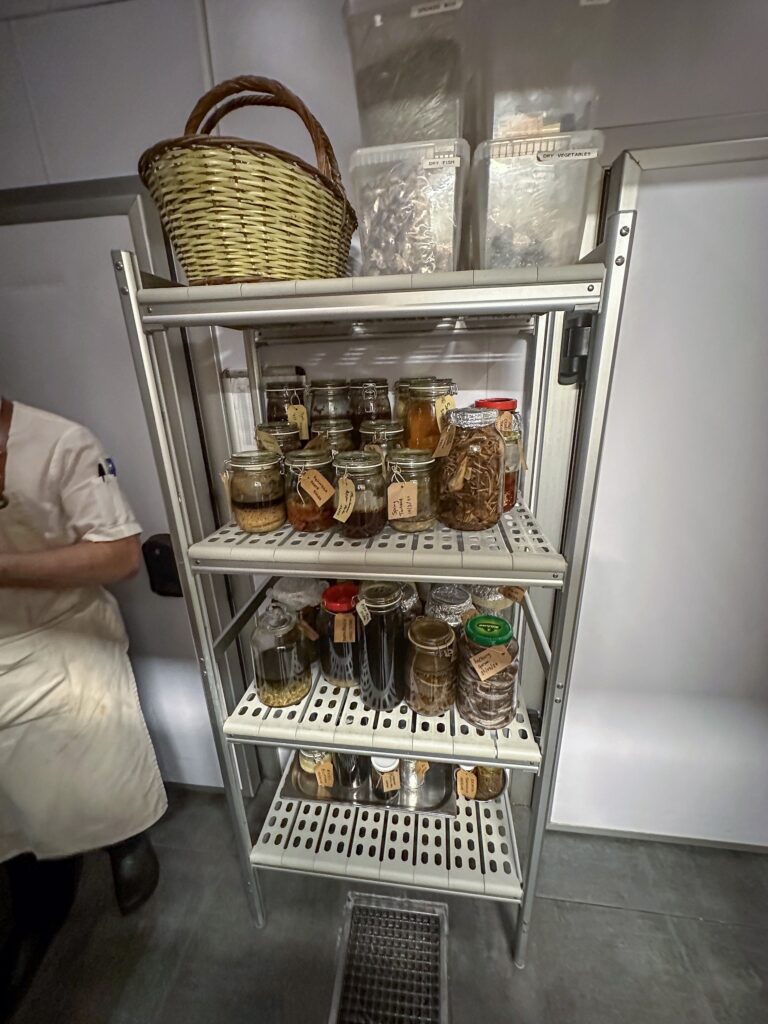 A rolling rack of labelled preserving jars and bottles in the kitchen, a wicker basket resting on top