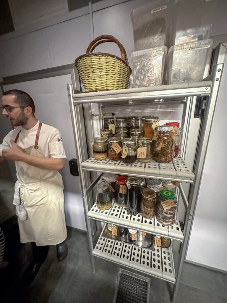 A cook standing beside the rolling rack of labelled ferments and preserves in the kitchen