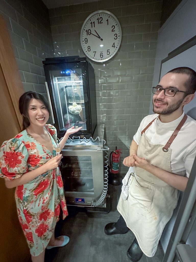 A cook and a diner in the kitchen beside the glass-fronted ageing cabinet, a wall clock behind them