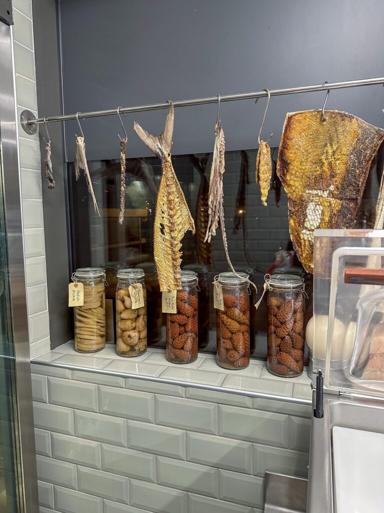 Whole fish hung to dry on a rail beside glass jars of preserved figs and dried chillies