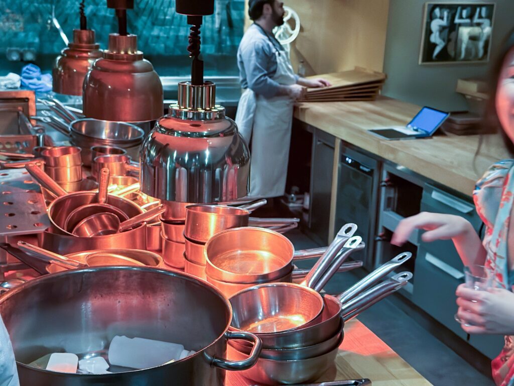 Copper pans stacked on the kitchen pass under brass heat lamps, a cook working at the counter behind