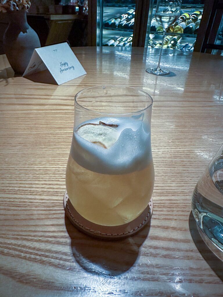 A tall glass of cloudy amber cocktail under a foam cap dusted with cinnamon, on a leather coaster