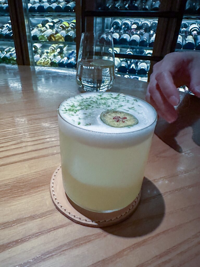 A pale green cocktail under a foam cap scattered with herbs and set with a dried lime wheel