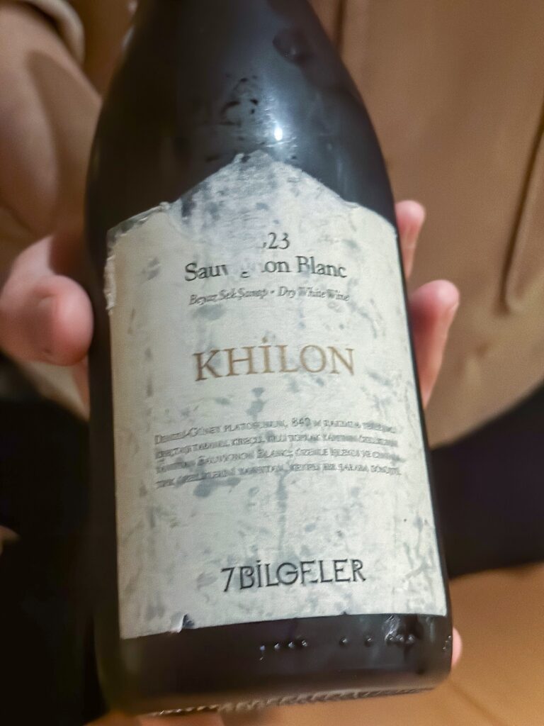 A hand holding a bottle of 7Bilgeler Khilon Sauvignon Blanc, its paper label torn and weathered