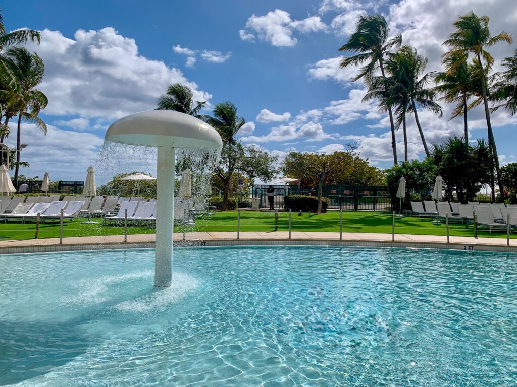 Mushroom fountain spilling into the shallow end of the family pool, ringed by palms and empty loungers