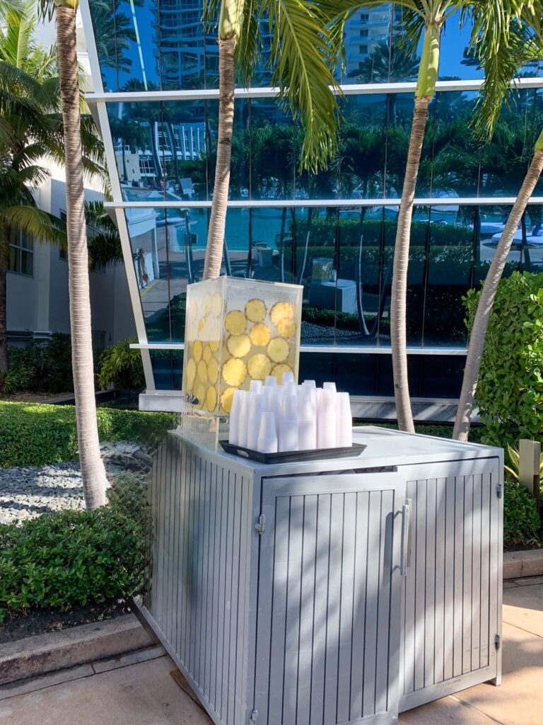 Glass dispenser of lemon infused water and stacked cups on a cart beside the palm shaded walkway