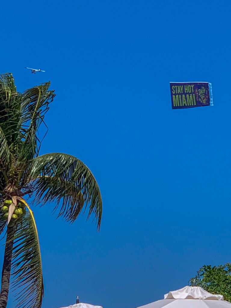Banner plane trailing a Stay Hot Miami sign above a coconut palm and pool umbrellas