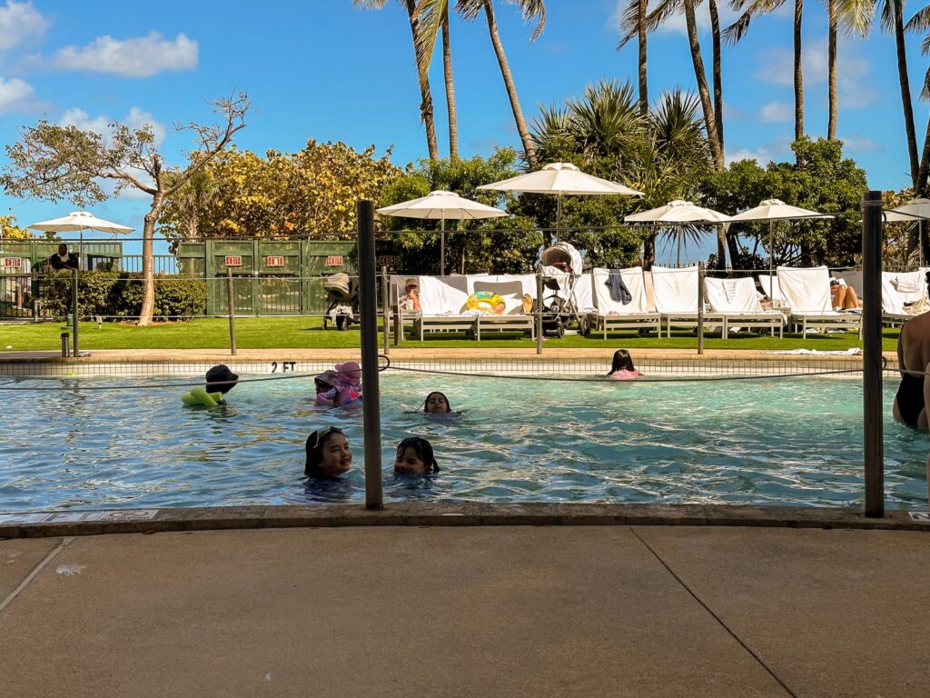Children swimming in the shallow family pool with the lawn, umbrellas and loungers beyond