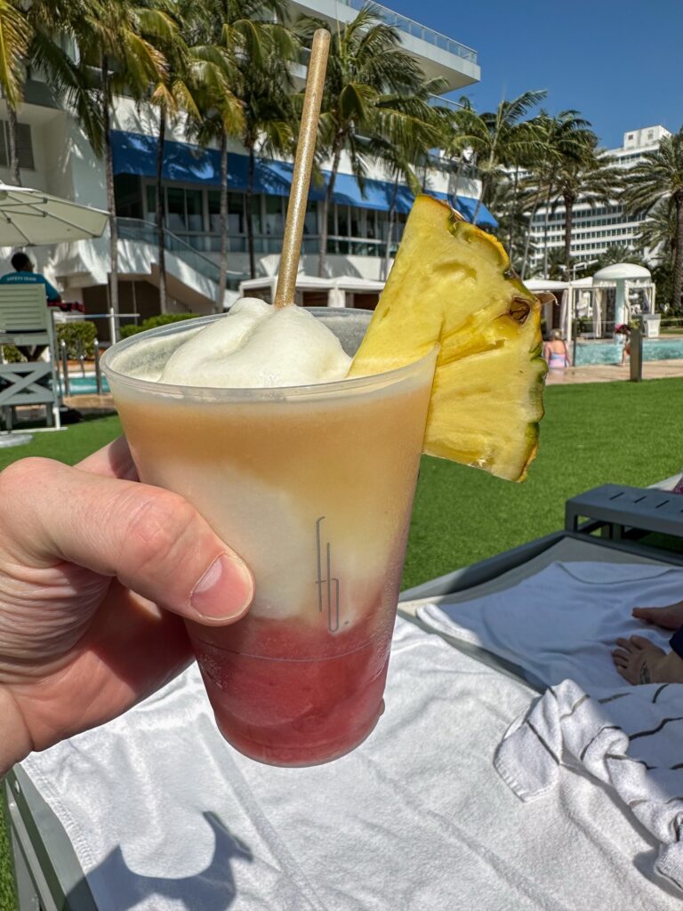 Layered frozen rum cocktail with a pineapple wedge held up in front of the pool lawn