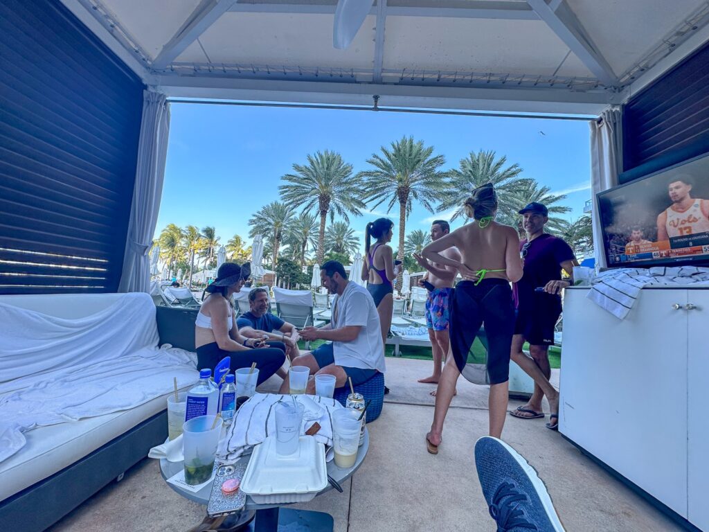 Inside a beach cabana with daybed, drinks table and television, looking out to the palms