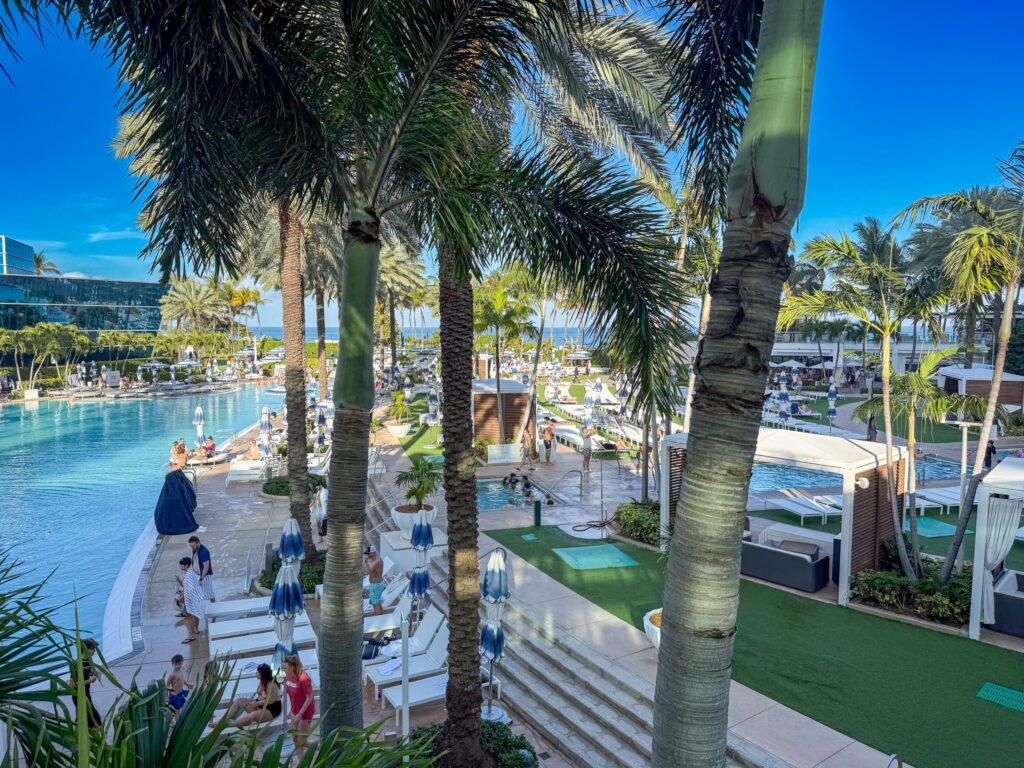 Palm trunks framing the pool deck, cabanas and turf lawn with the ocean beyond