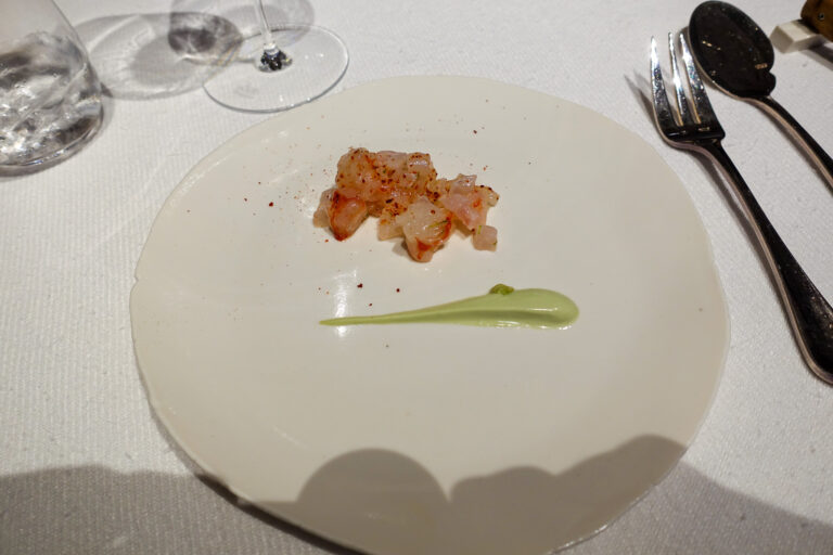 A small mound of diced pink seafood on a wide white plate beside a streak of pale green puree