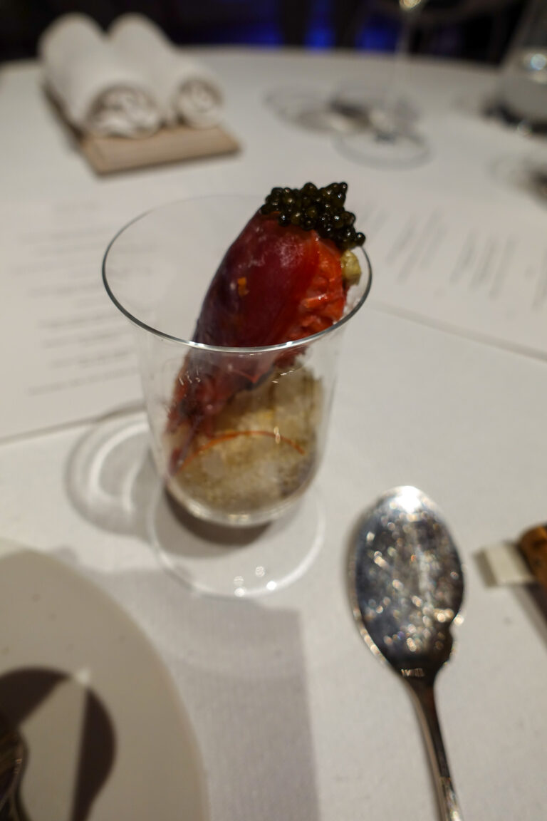 A whole red pepper crowned with caviar standing in a glass, the printed menu and a spoon alongside