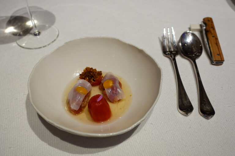Translucent slices and a peeled cherry tomato in a shallow pool of broth, set in a wide white bowl