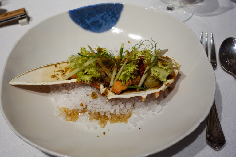 A dressed shellfish served in its shell on crushed ice, topped with herbs and shaved vegetables