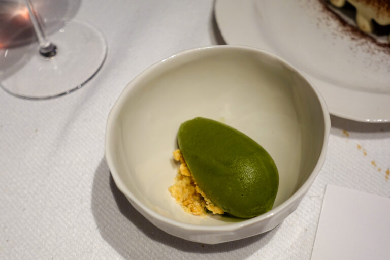 A green quenelle resting on a crumb base in a small white bowl