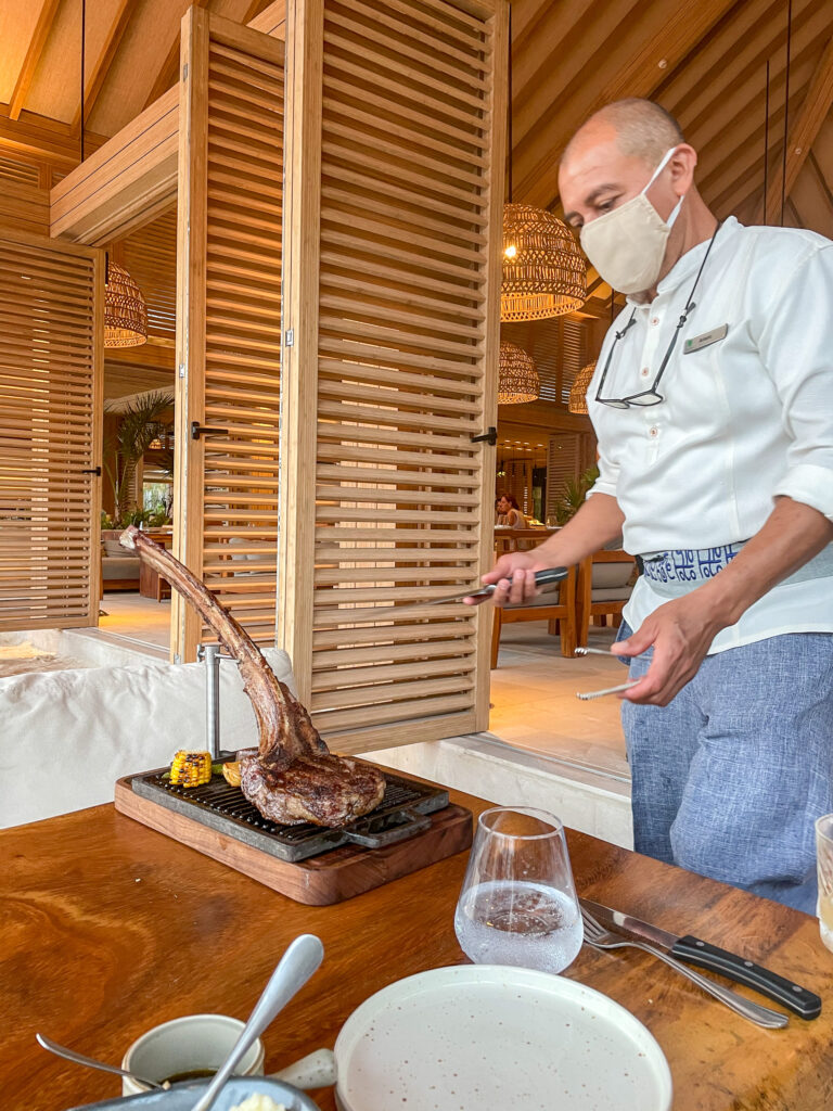 A chef carving the steak at the table beside a glass of wine