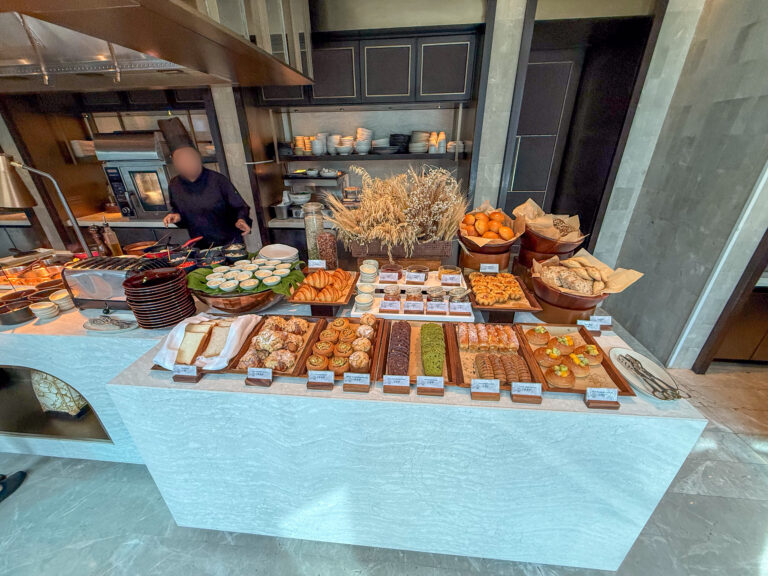 A long stone table laid with pastries and breads, a chef working at the range behind