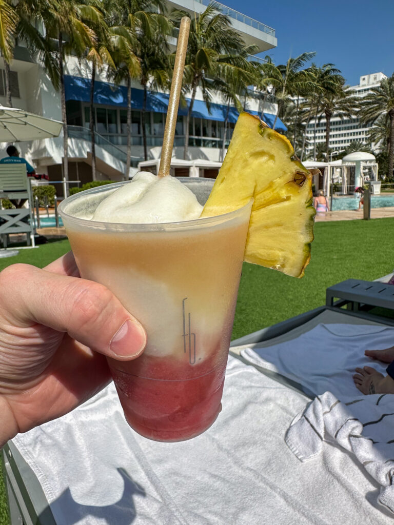 Layered frozen cocktail with a pineapple wedge held up in front of the pool lawn